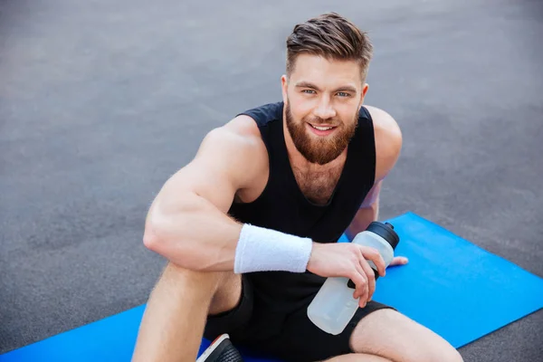 Lächelnder junger Sportler mit Flasche Wasser entspannt sich im Freien — Stockfoto