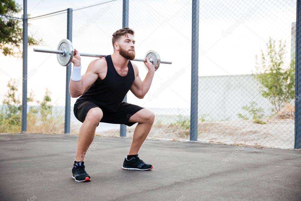 Concentrated bearded sports man doing squatting exercises with barbell