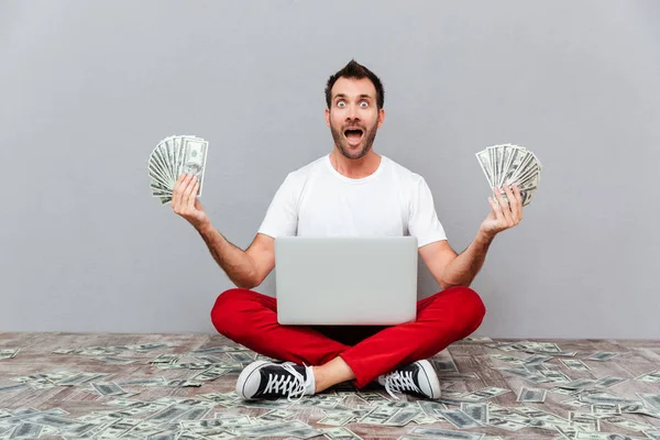 Muž sedí na podlaze s notebookem a drží peníze — Stock fotografie