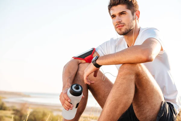 Красивый молодой спортсмен сидит и держит бутылку воды на открытом воздухе — стоковое фото