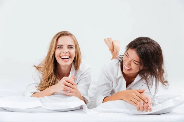 Due donne ridenti sdraiate insieme sul letto con lenzuola bianche — Foto Stock