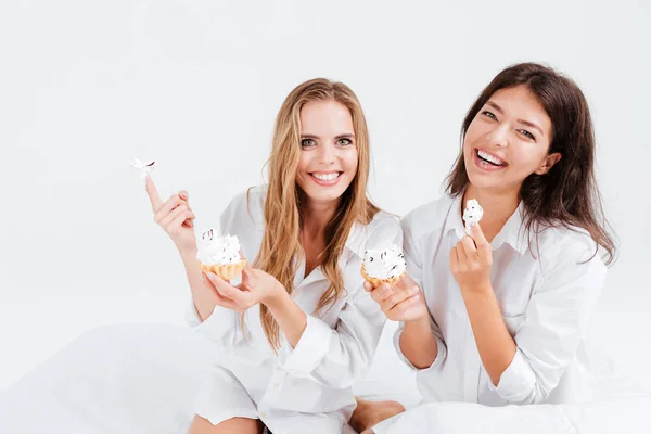 Dwie piękne kobiety wesoły jedzenia słodkich babeczek w łóżku — Zdjęcie stockowe