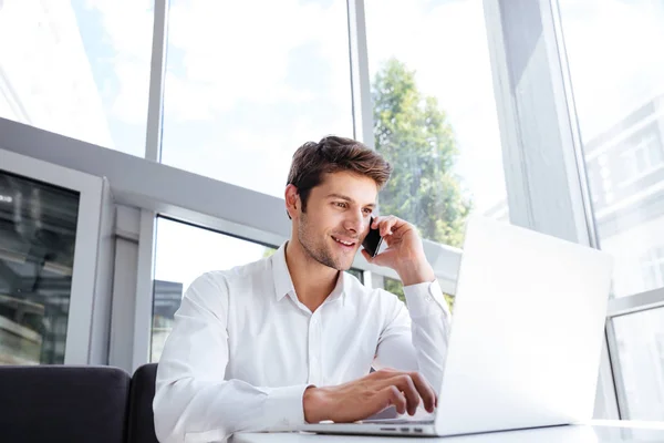 Feliz joven hombre de negocios hablando en el teléfono celular y utilizando el ordenador portátil — Foto de Stock