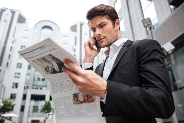 Joven empresario concentrado hablando por celular y leyendo el periódico — Foto de Stock