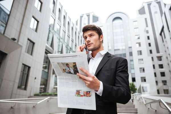 Joven empresario pensativo hablando por teléfono móvil y leyendo el periódico — Foto de Stock