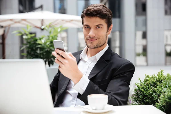 Hombre de negocios feliz utilizando el ordenador portátil y teléfono inteligente en la cafetería al aire libre —  Fotos de Stock