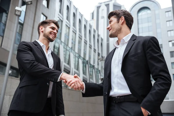 Dos empresarios felices de pie y estrechando las manos cerca del centro de negocios — Foto de Stock