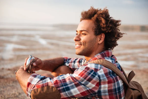 Usmívající se muž s batohem pití z placatky na pláži — Stock fotografie