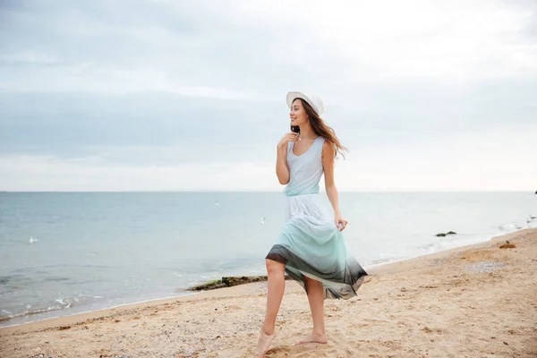 Vrouw in witte hoed en jurk wandelen op het strand — Stockfoto