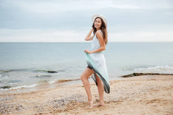 Счастливая женщина в шляпе и платье, стоящие на пляже — стоковое фото