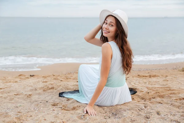 微笑的帽子和衣服坐在沙滩上的女人 — 图库照片