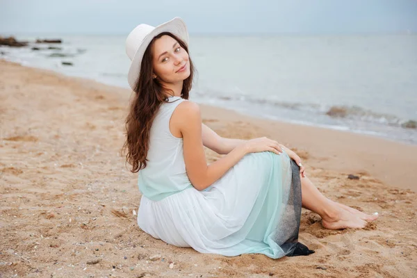 Frau mit Hut und Kleid sitzt barfuß am Strand — Stockfoto