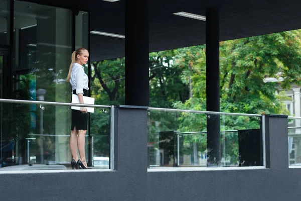 Joven mujer de negocios sosteniendo portátil de pie en el edificio de vidrio —  Fotos de Stock