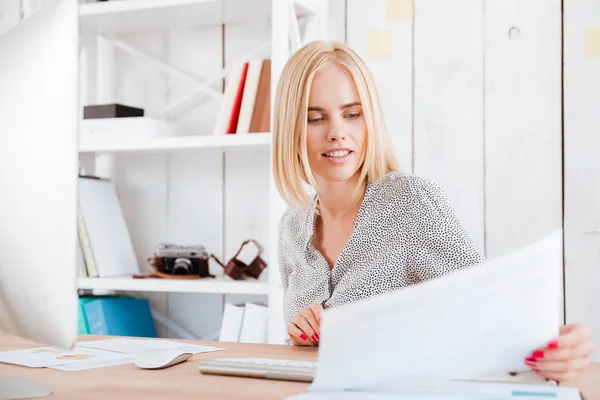 Koncentrovaná světlovlasá žena, zatímco sedí na pracovišti při pohledu přes dokumenty — Stock fotografie