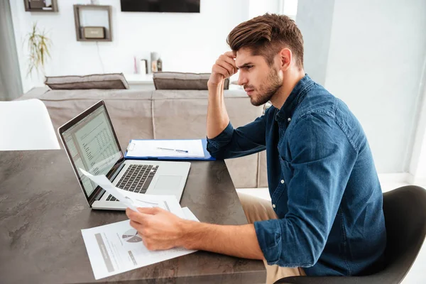 Concentrated man using laptop and looking on papers — Stock Photo, Image