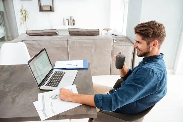 Joven alegre bebiendo café mientras está sentado cerca de la computadora portátil — Foto de Stock