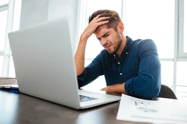 Confused young man sitting near laptop and holding head — Stock Photo, Image