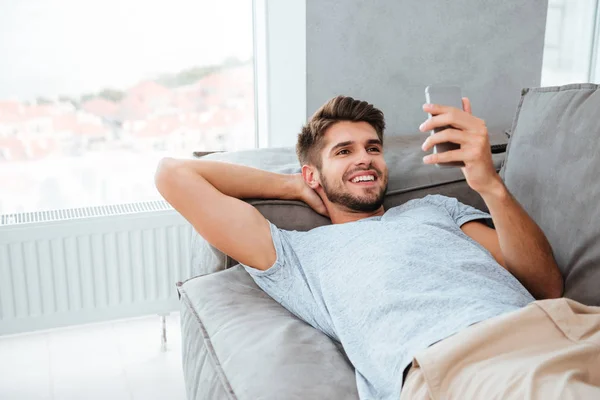 Šťastný mladý muž leží na posteli a při pohledu na telefon — Stock fotografie