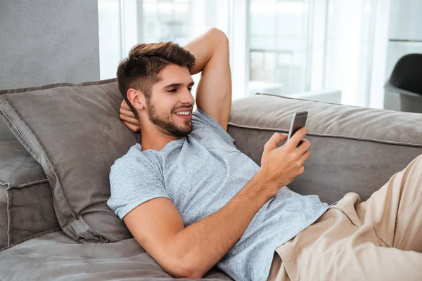 Šťastný mladý muž leží na pohovce a chatování na telefonu — Stock fotografie