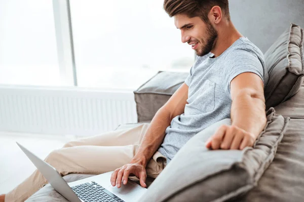 Joyeux jeune homme assis sur le canapé et travaillant par ordinateur portable — Photo