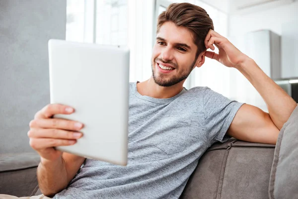 Mladý, šťastný muž sedí na pohovce a při pohledu na tabletu — Stock fotografie