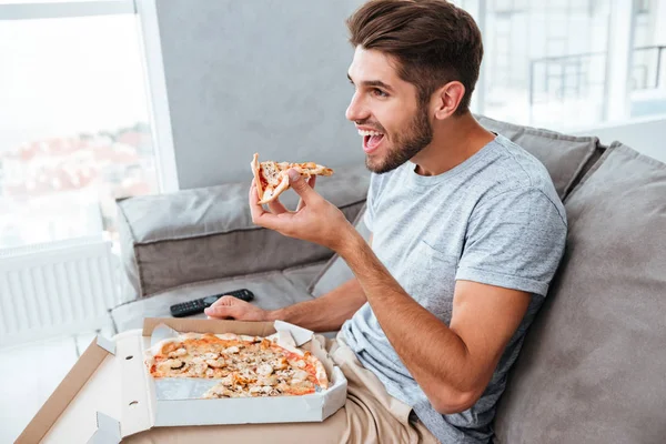 Mand spiser pizza, mens du sidder på sofaen og ser tv - Stock-foto