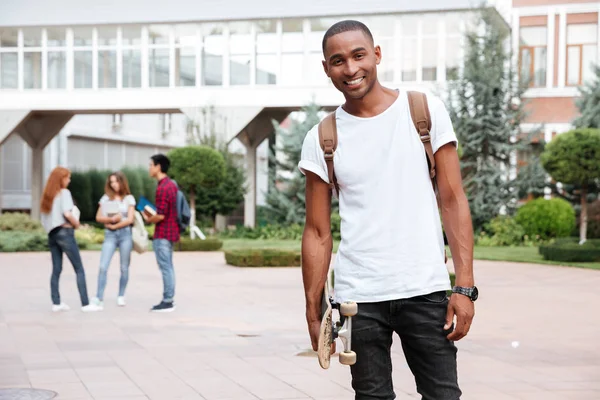 Alegre estudante homem africano com mochila segurando skate ao ar livre — Fotografia de Stock