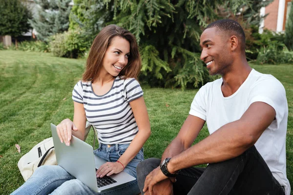 Feliz casal multiétnico sentado e usando laptop no gramado ao ar livre — Fotografia de Stock