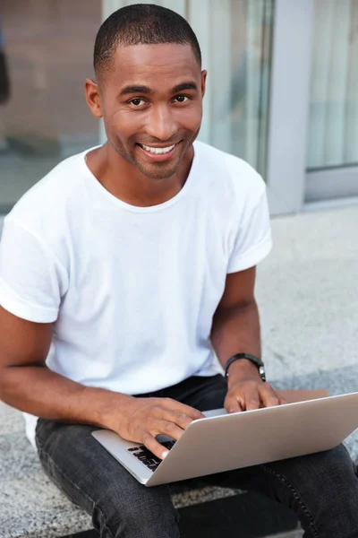 Vrolijke Afrikaanse jonge man zitten en met laptop buiten — Stockfoto