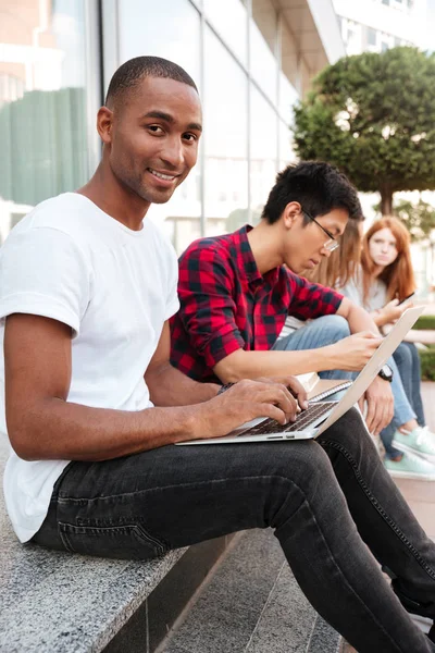 Sorrindo homem africano sentado com amigos e usando laptop ao ar livre — Fotografia de Stock