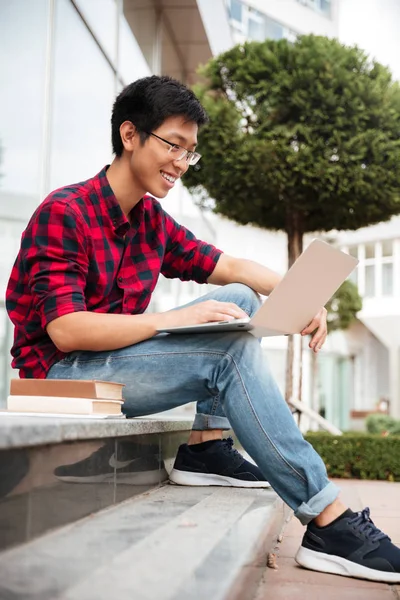 Vrolijke Aziatische jonge man zitten en met laptop buiten — Stockfoto