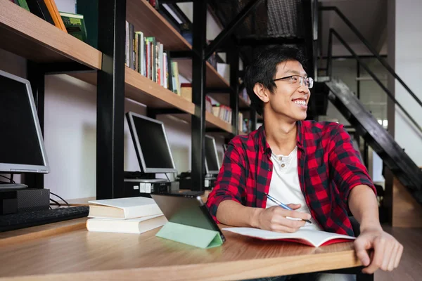 Щасливий азіатський студент чоловічої статі, використовуючи планшет у бібліотеці та написанні — стокове фото