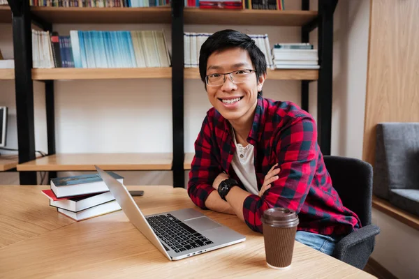 Kütüphanede dizüstü kullanarak ve kahve içme Asyalı adam — Stok fotoğraf
