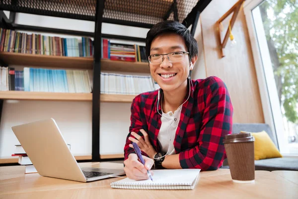 Asiatico maschio utilizzando laptop in biblioteca mentre scrivere a notebook — Foto Stock