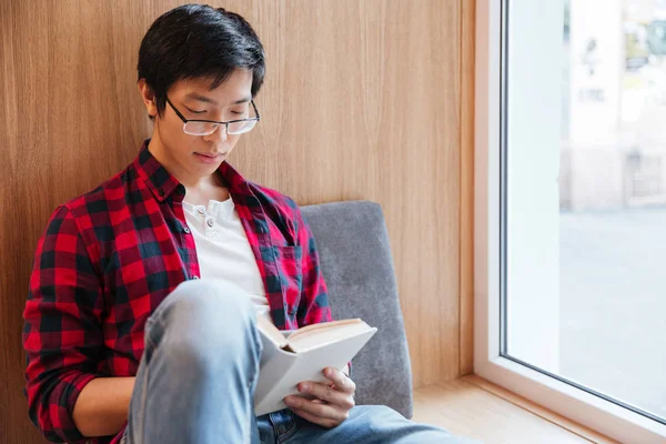 Azjatycki student czytania książki w bibliotece Uniwersyteckiej, siedząc na kanapie — Zdjęcie stockowe