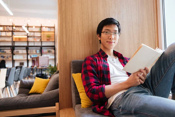Asiatische Studentin liest Buch in Universitätsbibliothek auf Sofa sitzend — Stockfoto