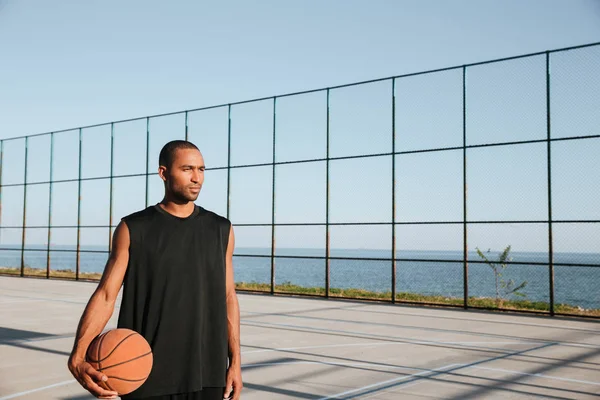 Yuong afro-americano desportista segurando basquete e olhando para longe — Fotografia de Stock
