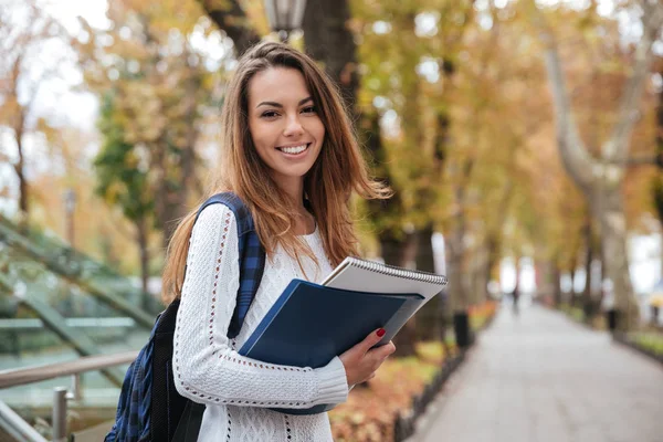 Cheerful young woman with backpack and notebooks standing in park — Stock Photo, Image