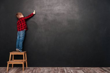 Child standing on stool over chalkboard and drawing at board clipart