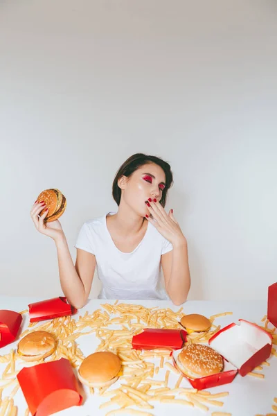 Mujer joven cansada comiendo hamburguesa y bostezando en la mesa — Foto de Stock