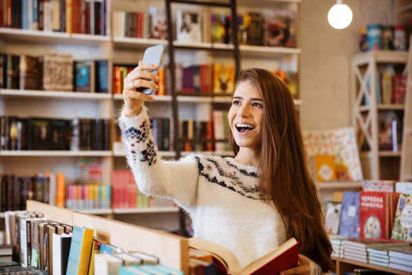 Lächelndes glückliches Mädchen macht Selfie, während es in der Bibliothek sitzt — Stockfoto