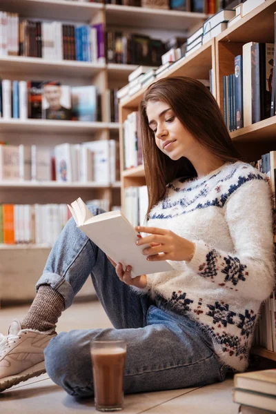 Hezká student čtení knihy na podlaze knihovny na univerzitě — Stock fotografie