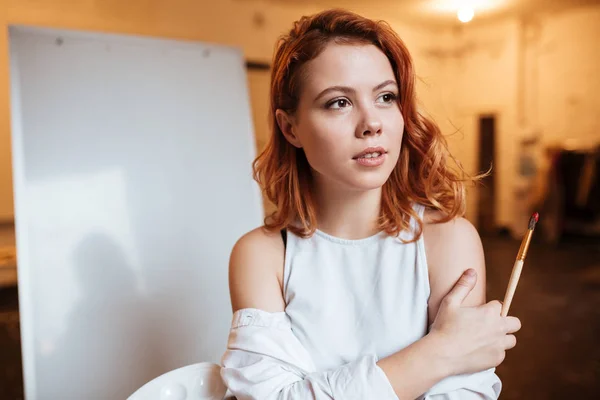 Concentrated lady painter with red hair standing over blank canvas