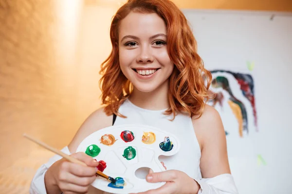 Senhora pintora sorridente com cabelo vermelho em pé sobre tela em branco — Fotografia de Stock