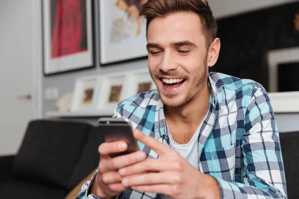 Mladý se smíchem muž sedí v domácnosti a při pohledu na telefon — Stock fotografie