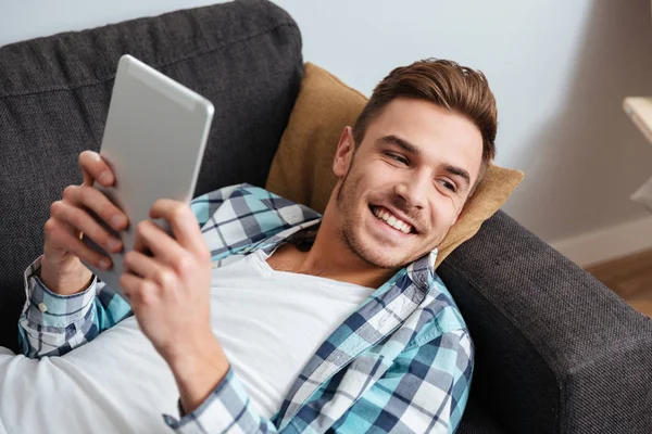 Hombre de la cerda sonriente se encuentra en el sofá y el uso de la computadora tableta — Foto de Stock