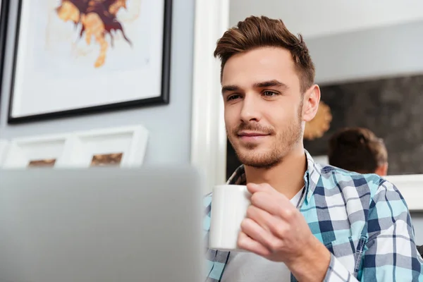Hombre joven feliz usando el ordenador portátil mientras bebe café —  Fotos de Stock