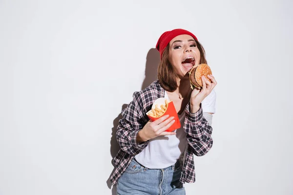 Hambrienta joven emocional sosteniendo papas fritas y hamburguesa —  Fotos de Stock