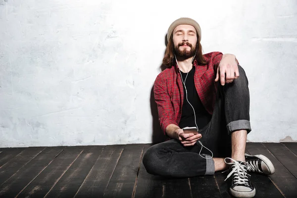 Веселий молодий бородатий хіпстер сидить на підлозі — стокове фото