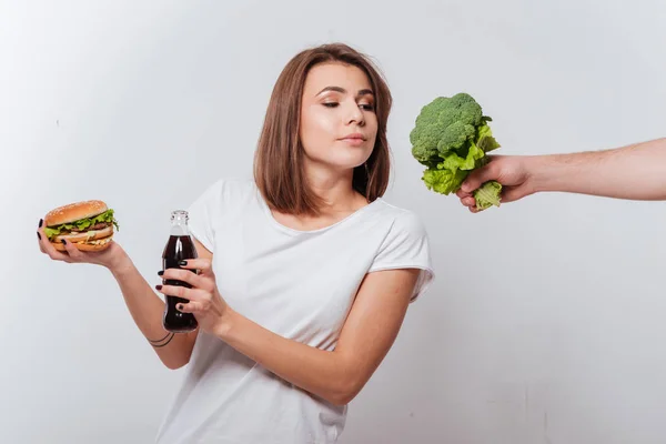 Hongerige jonge dame bedrijf fastfood en kijken broccoli — Stockfoto